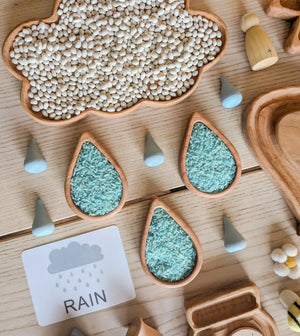 Cloud-shaped wooden sensory tray filled with white beans and raindrop-shaped trays with blue rice on wood table