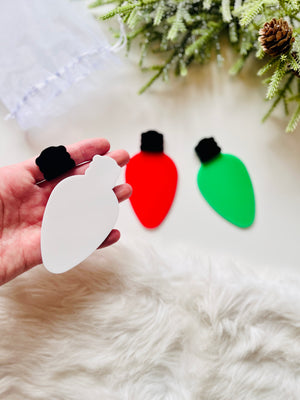 Hand holding white acrylic Christmas light bulb shape with red and green bulbs and pine branches in background
