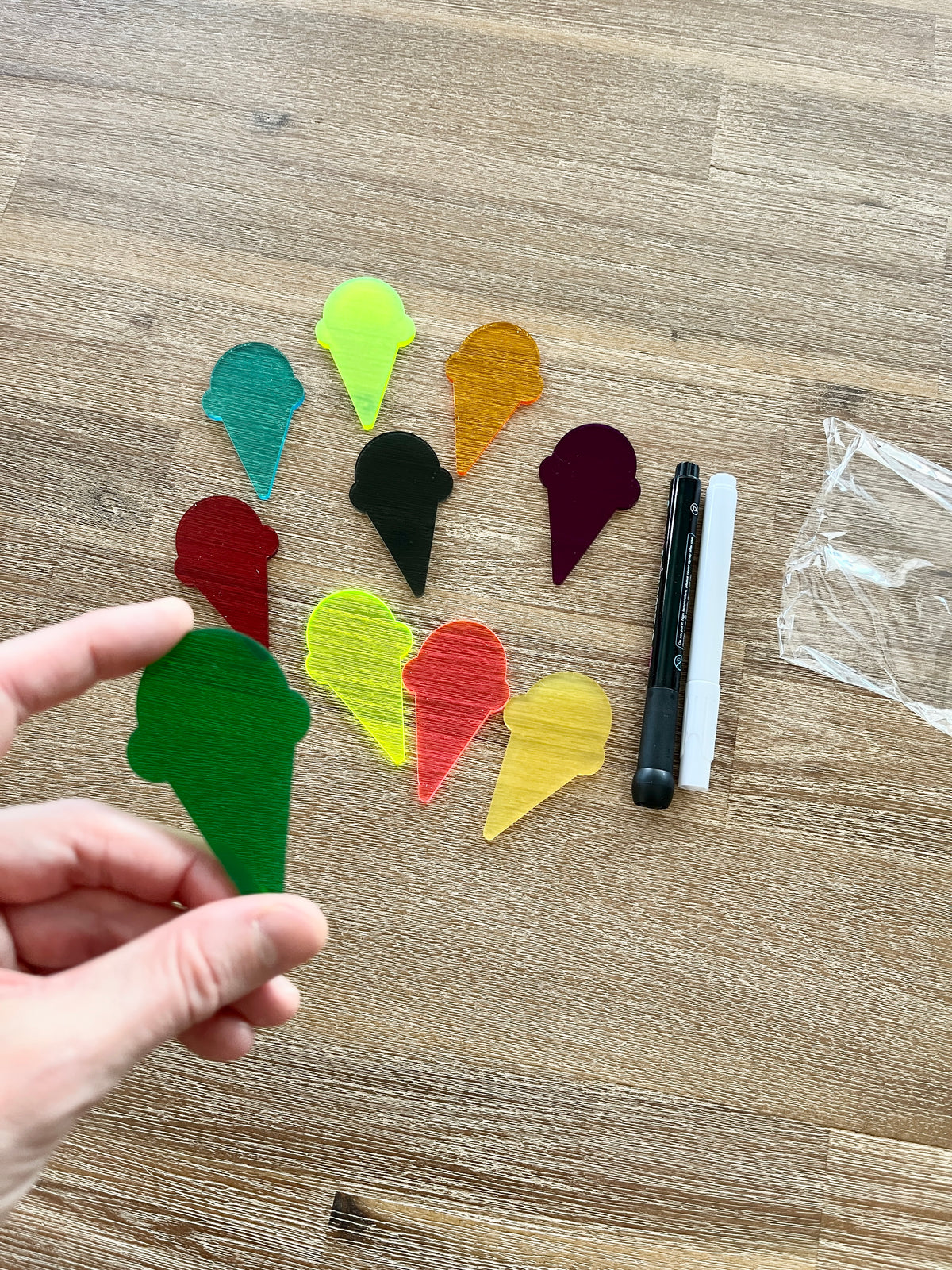 Hand holding a large translucent green ice cream cone piece from Little Dry Erasables - Translucent Ice Cream Cones collection, with colorful ice cream cone pieces and black and white dry erase markers on a wooden surface, featuring handcrafted homeschool education resources.