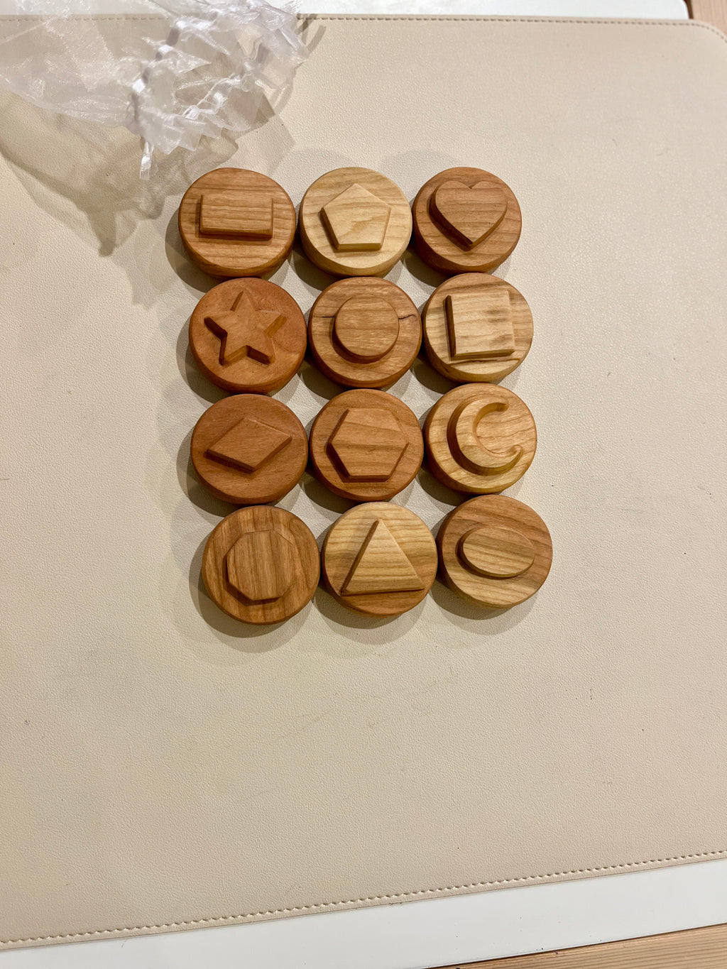 Twelve round wooden tokens featuring various raised geometric shapes arranged on a light surface with plastic wrapping nearby