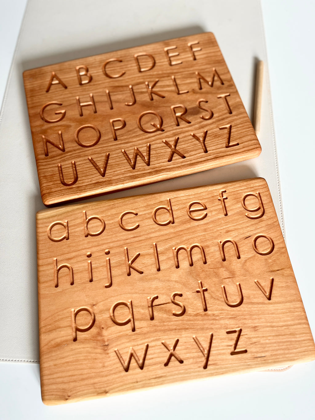 Two wooden alphabet tracing boards with carved uppercase and lowercase letters and a wooden stylus on white surface