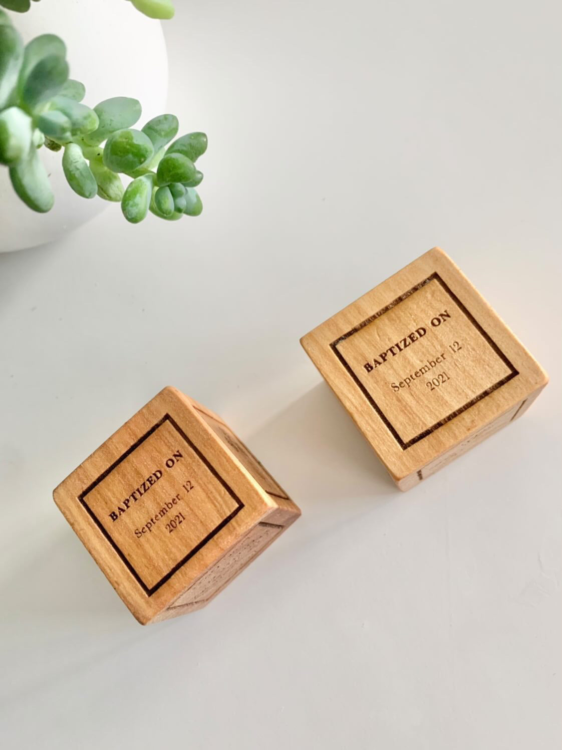 Two personalized wooden baptism blocks engraved with baptism date displayed on white surface next to green plant