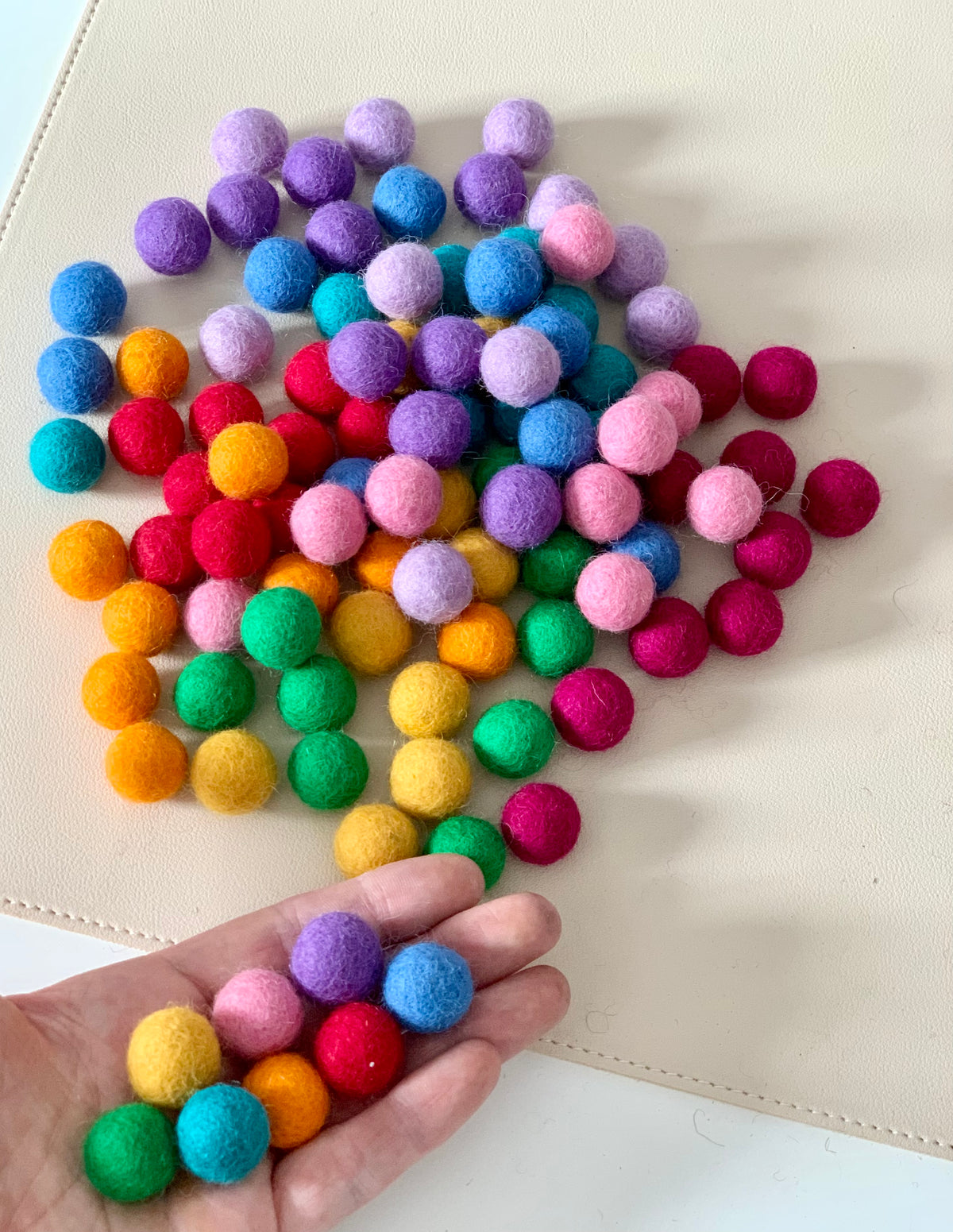 Colorful 1.5cm felt balls in a variety of rainbow colors held in hand and scattered on cream surface