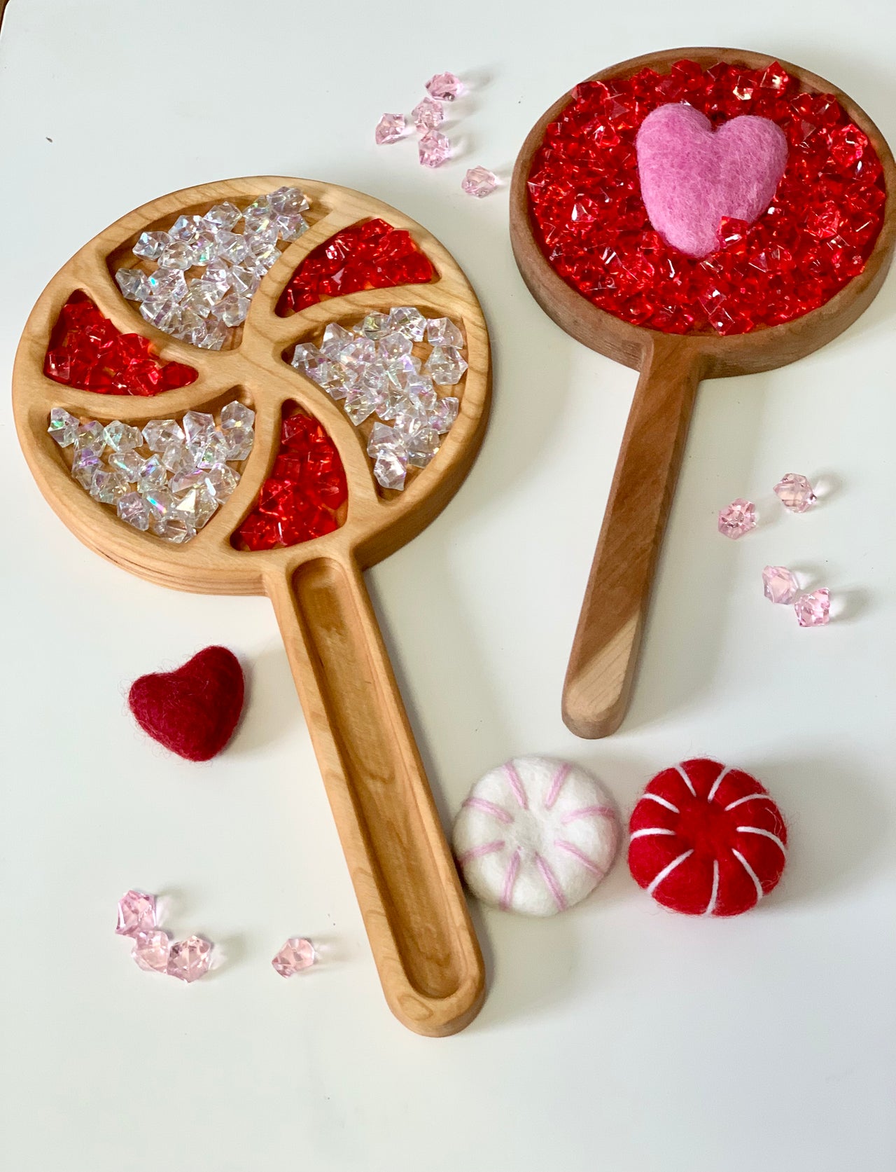 Two wooden handheld trays filled with red and clear acrylic gems and felt heart and peppermint shapes scattered nearby