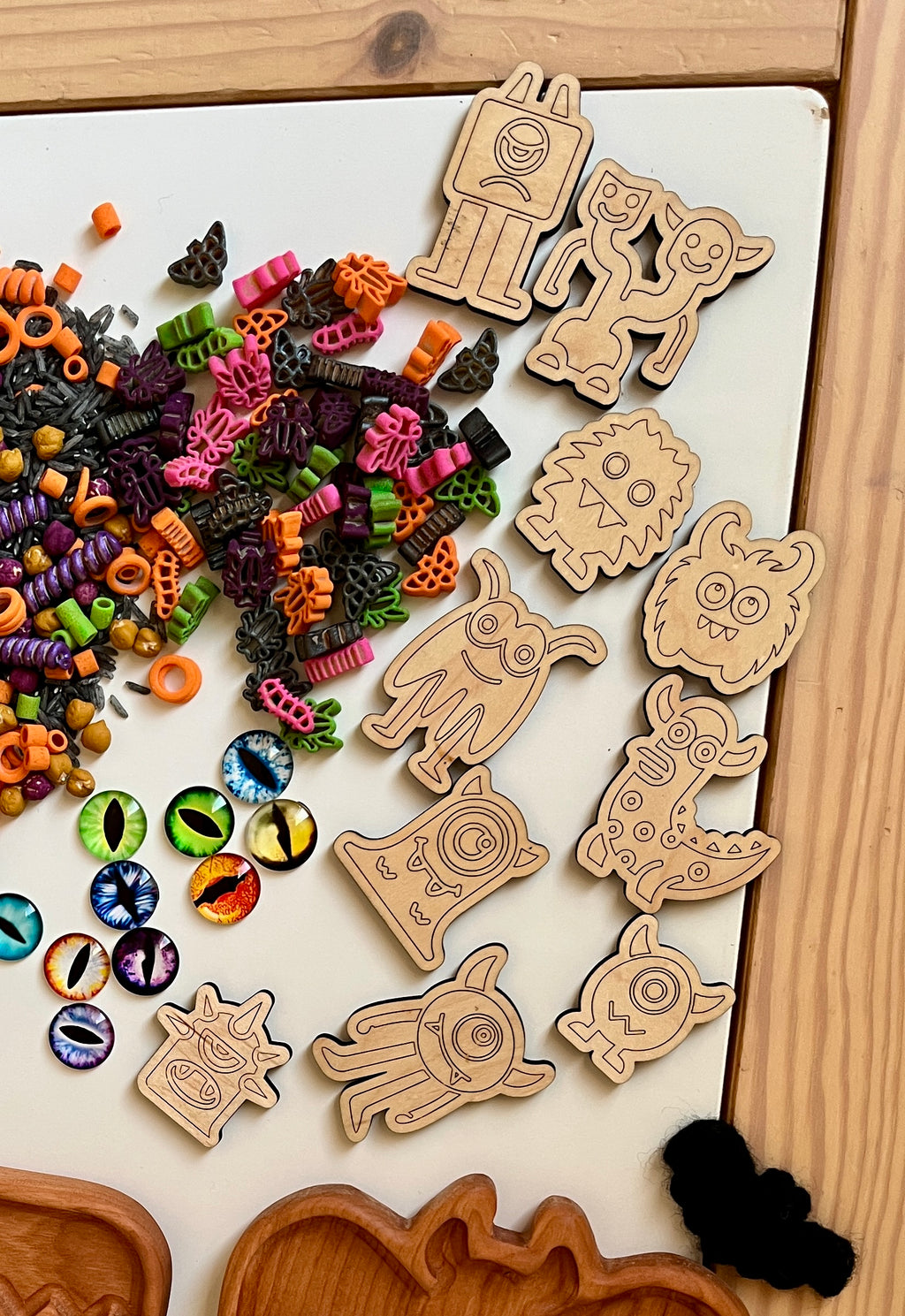 Wooden monster tokens with engraved faces, colorful Halloween pasta shapes, and decorative monster eye pieces on white surface