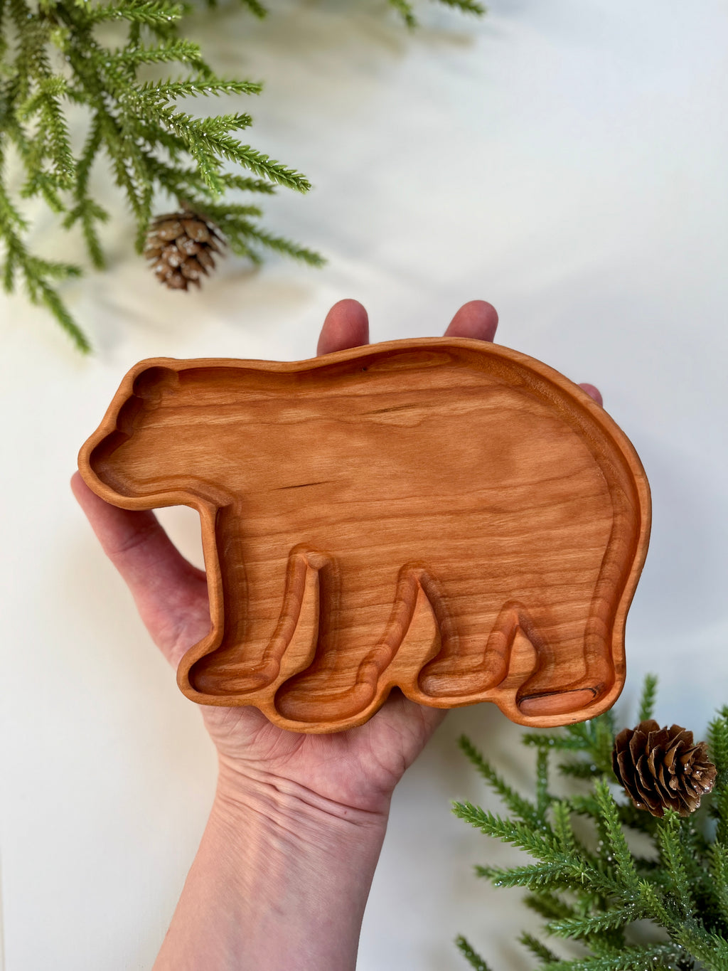 Hand holding a wooden tray carved in the shape of a bear with pine branches and pine cones nearby on white surface