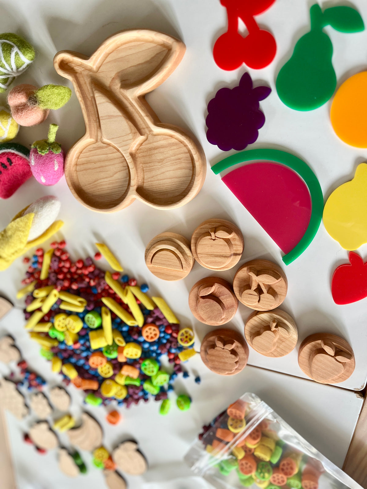 Wooden cherry-shaped tray surrounded by colorful felt fruits, wooden buttons, small beads, and craft supplies on white background