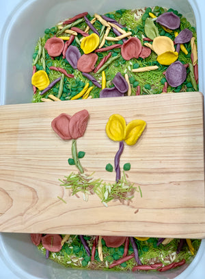 Colorful clay flower shapes arranged on a wooden board above mixed dried peas, rice, and pasta in a container