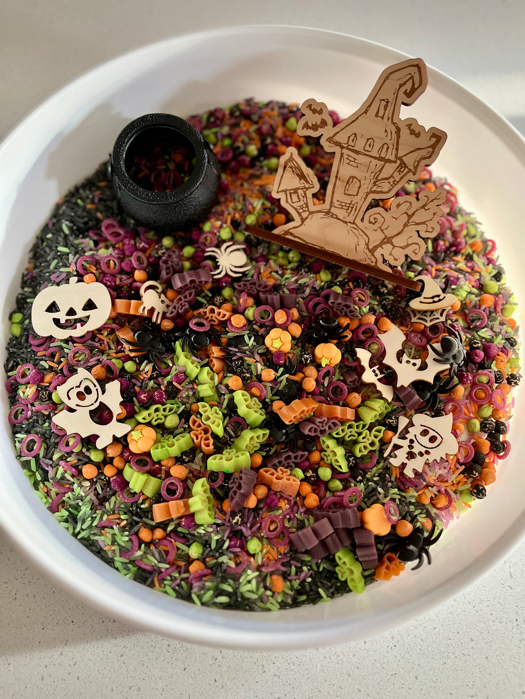 White bowl with multicolored Halloween-themed pasta shapes and wooden haunted house and spooky character decorations inside