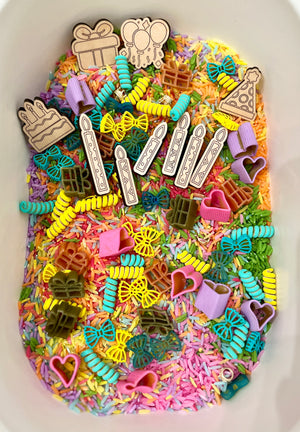 Colorful dyed rice filled with wooden birthday-themed tokens, candles, and pasta shaped like bows, hearts, and gifts