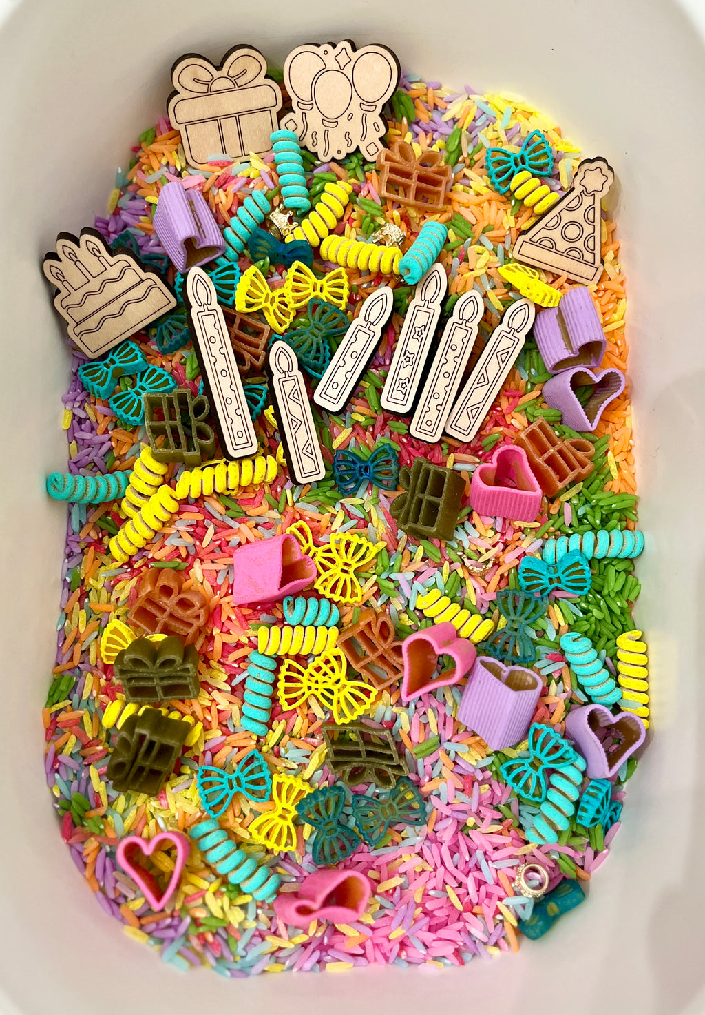 Colorful dyed rice filled with wooden birthday-themed tokens, candles, and pasta shaped like bows, hearts, and gifts