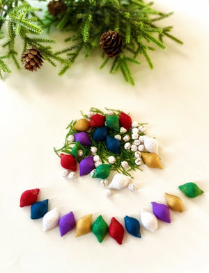 Assorted colorful textured fabric capsules and white berry clusters arranged on a light surface with pine branches and cones above