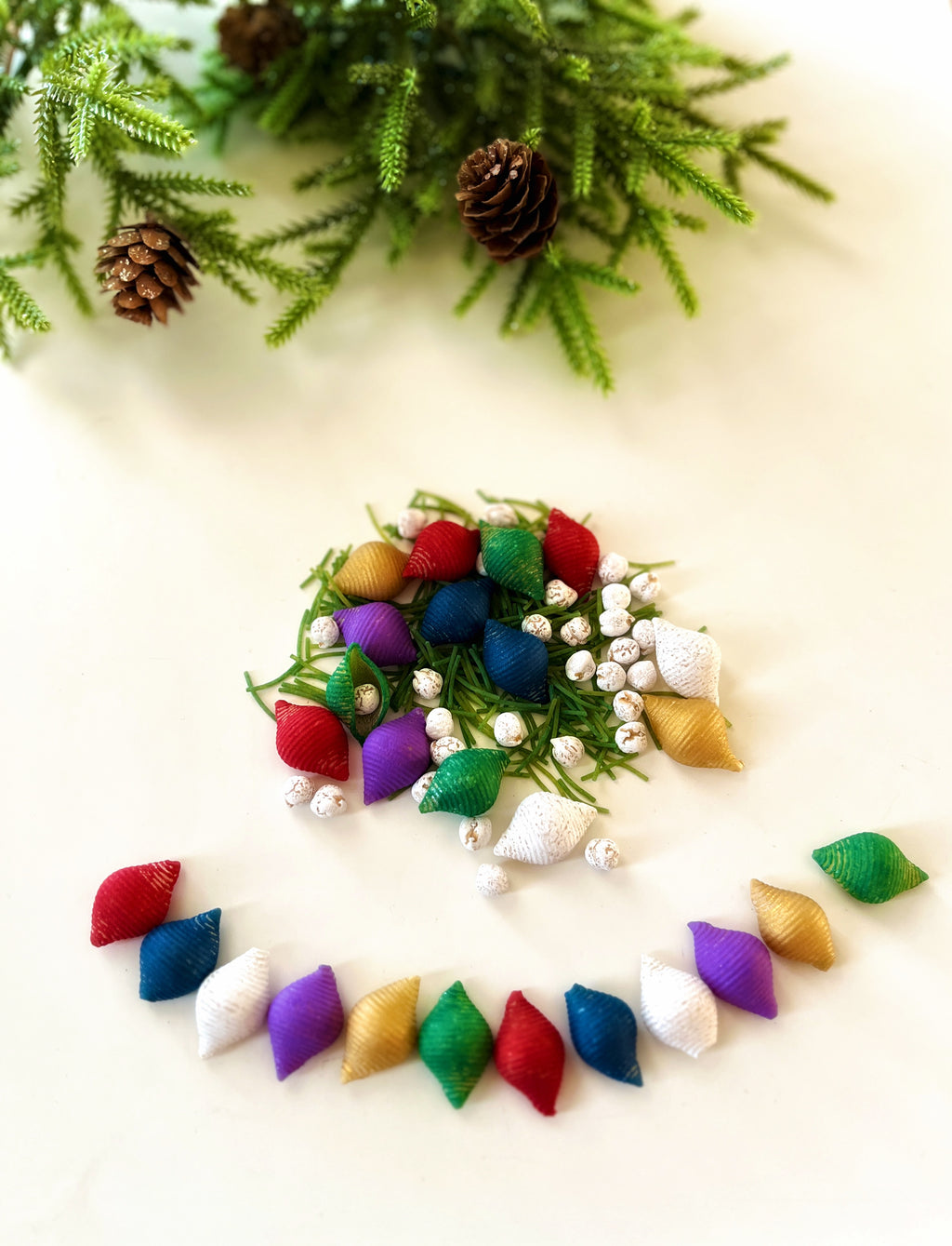 Assorted colorful textured fabric capsules and white berry clusters arranged on a light surface with pine branches and cones above