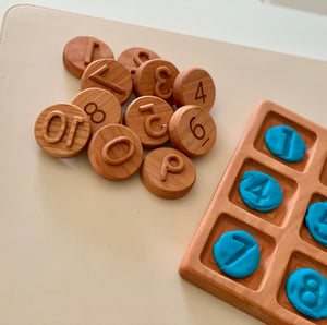 Round wooden tokens engraved with numbers placed beside a wooden grid tray holding blue number-shaped clay pieces