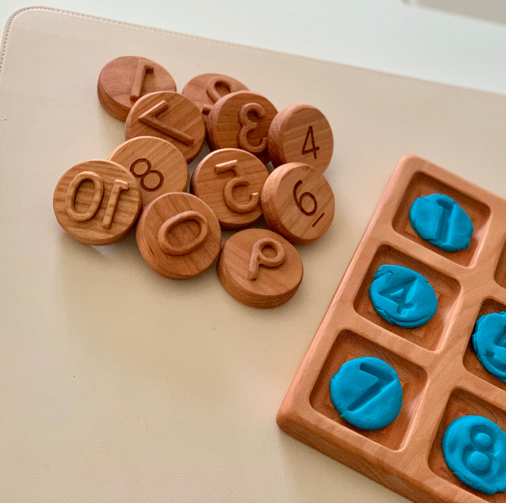 Round wooden tokens engraved with numbers placed beside a wooden grid tray holding blue number-shaped clay pieces