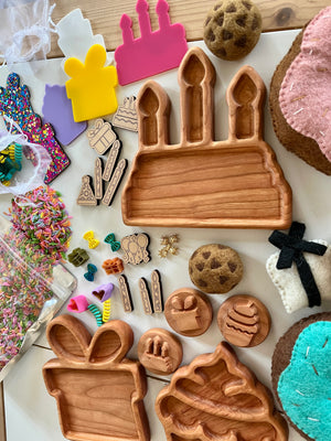 Wooden birthday-themed trays shaped like cake, gift, and cupcake surrounded by colorful craft supplies and felt cookies