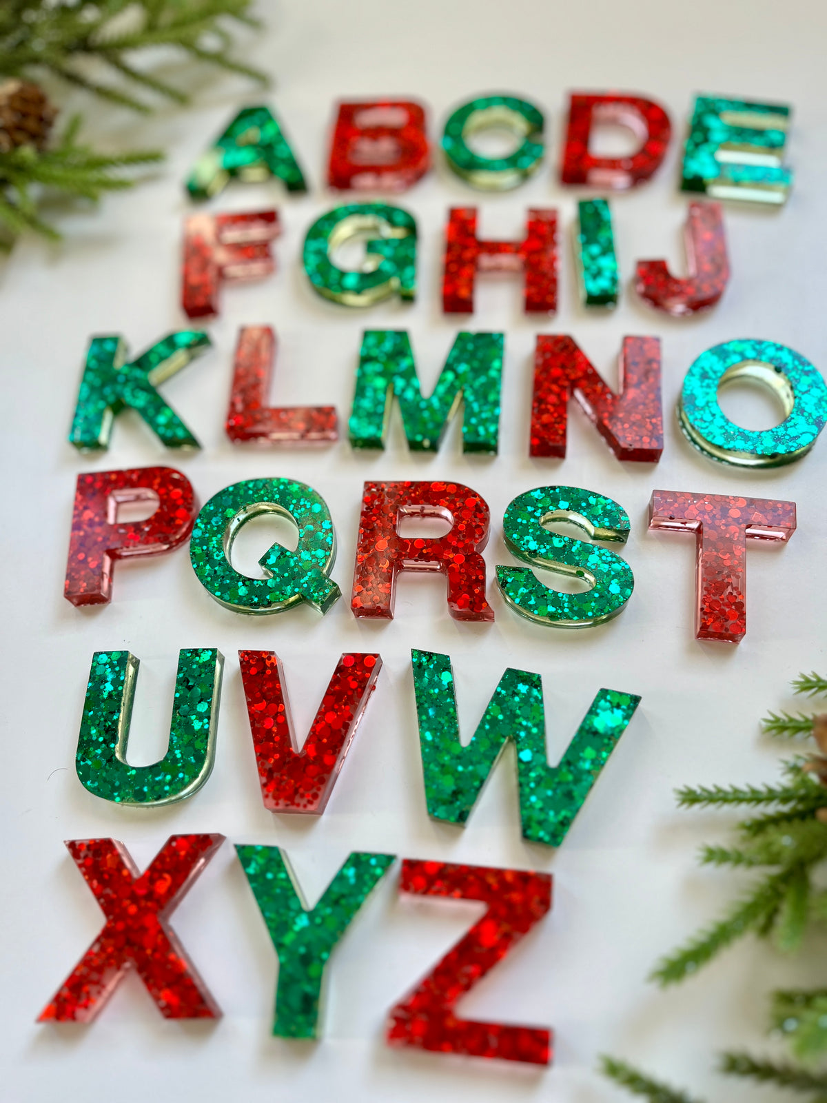 Glittery red and green resin letters arranged on white surface with pine branches in corners