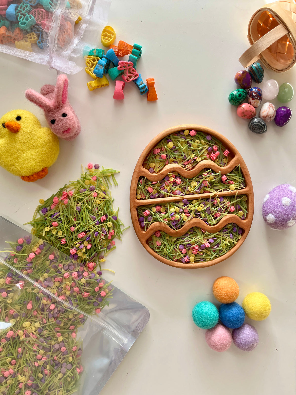 Wooden egg-shaped tray with dried green grass and pink flowers surrounded by colorful felt balls and decorative stones