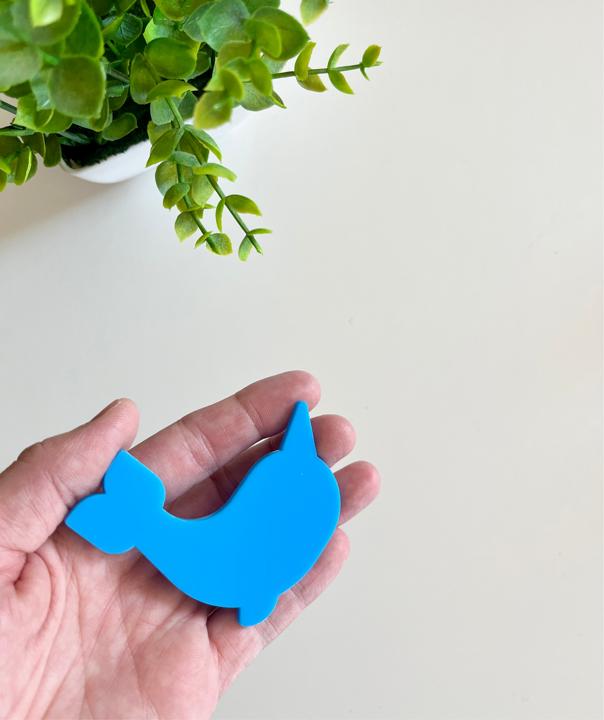 Hand holding a smooth blue whale-shaped cutout with a green leafy plant in a white pot nearby on a light surface