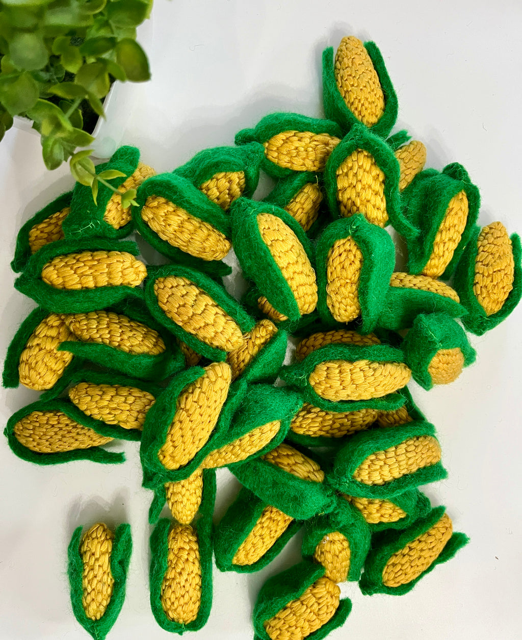 Pile of handmade knitted yellow corn cobs with green husks arranged on white surface near small green plant