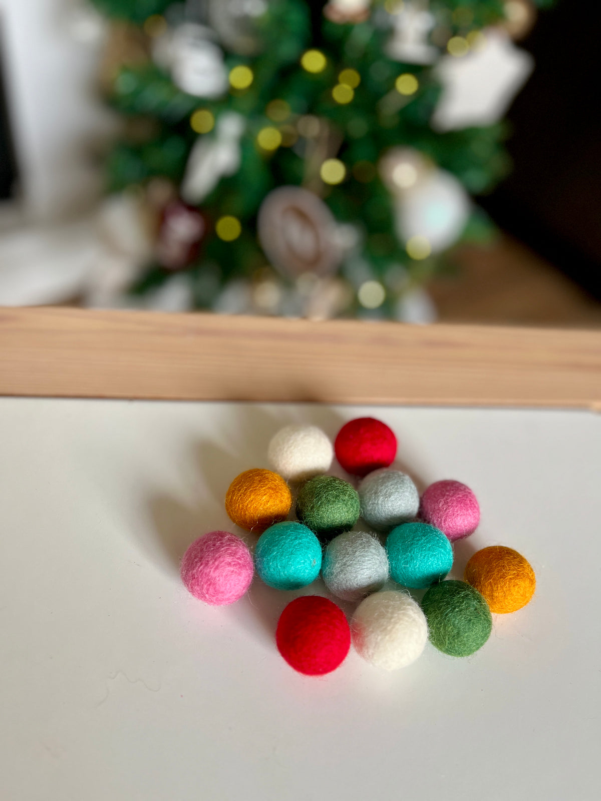 Multicolored felt balls in pink, red, green, orange, white, and blue arranged on a white surface with blurred tree background