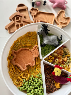 White divided tray with green, yellow, and brown pasta, wooden pig scoop, felt horse and rooster, and wooden farm shape cookie cutters nearby