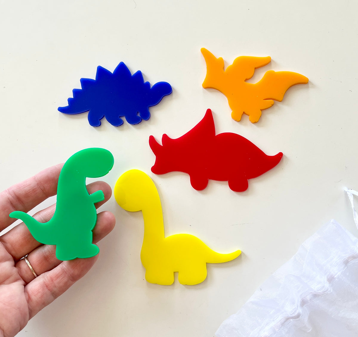 Bright green, yellow, blue, red, and orange acrylic dinosaur shapes on a white surface with a hand holding the green one