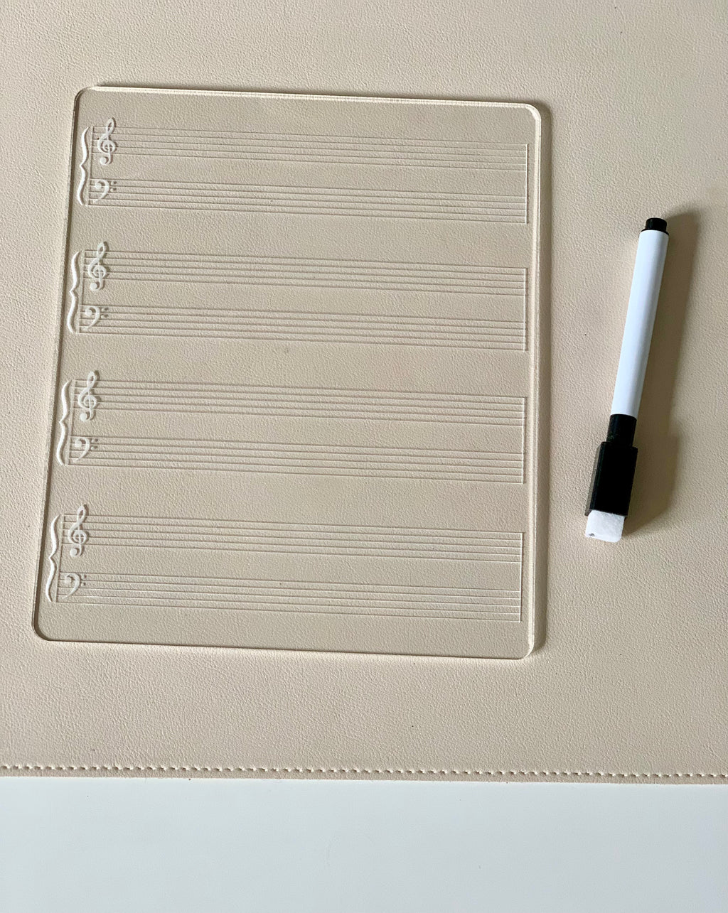 Clear music staff writing board with treble and bass clefs next to a black and white dry erase marker on beige surface