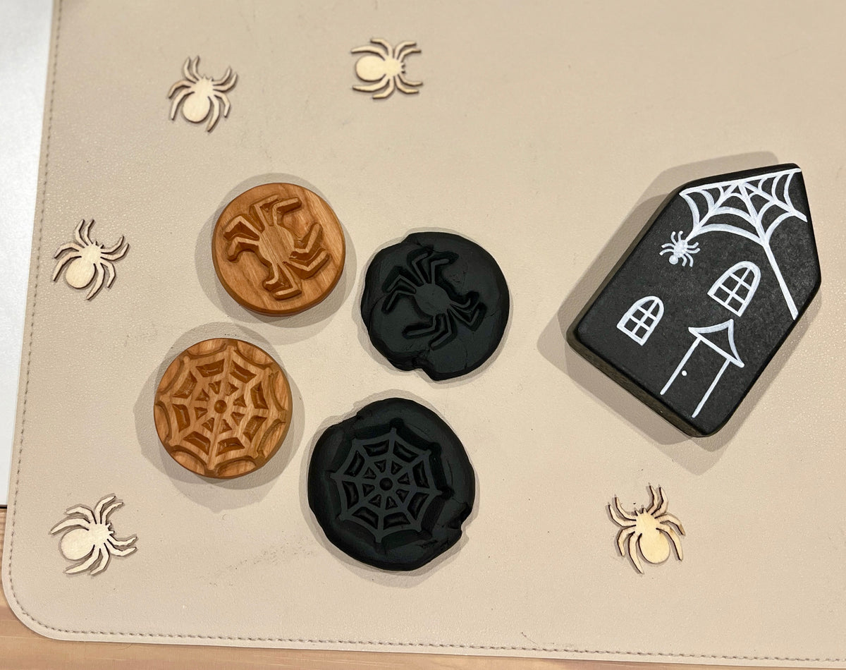 Wooden and black carved tokens with spider and web designs, small wooden spider cutouts, and a black house with a spiderweb decoration