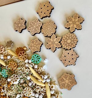 Laser-cut wooden snowflake shapes arranged above mixed sensory materials with pasta and small white beads on white surface