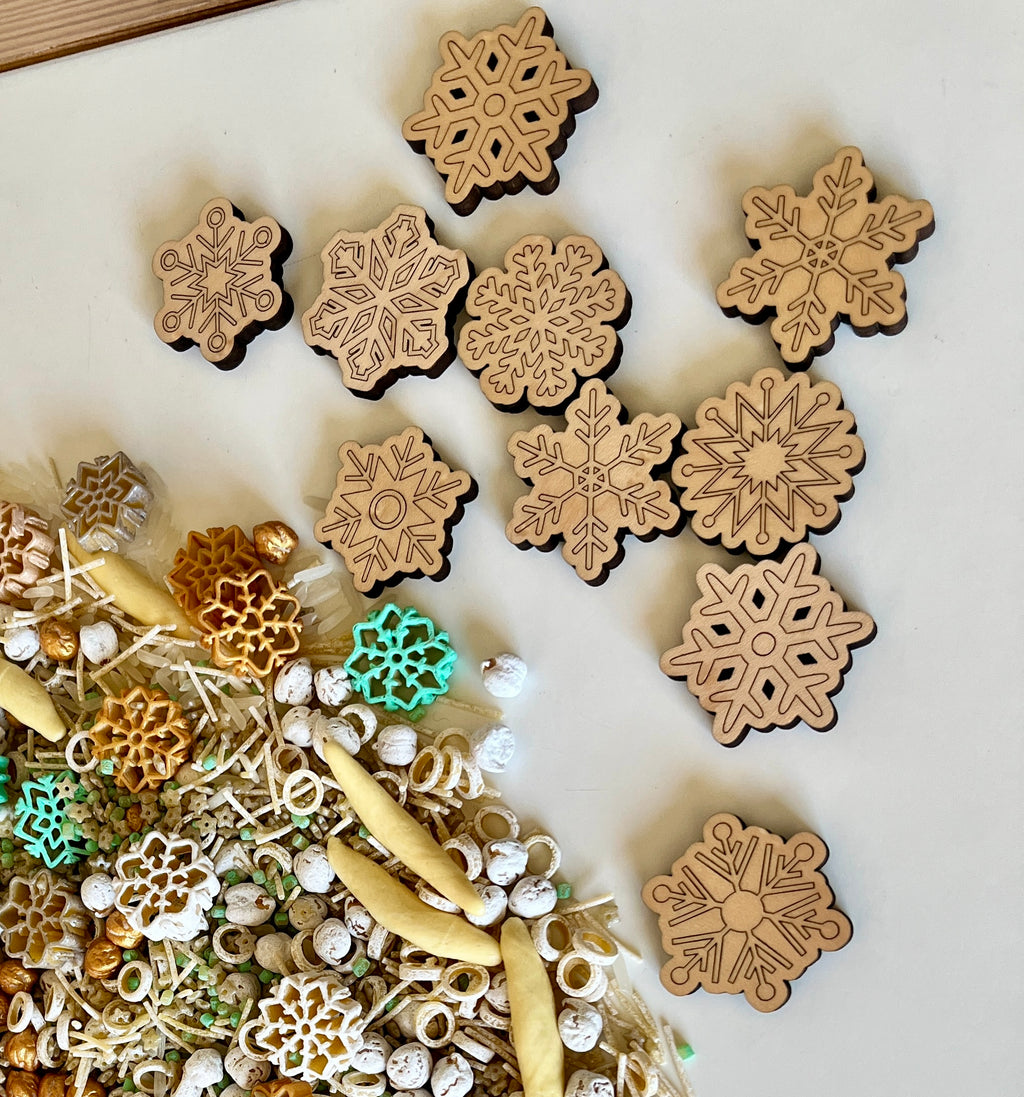 Laser-cut wooden snowflake shapes arranged above mixed sensory materials with pasta and small white beads on white surface