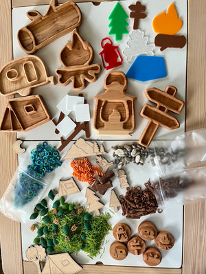 Wooden camping-themed trays with tent, camper, fire, lantern, trees, signs, play pieces, beads, and natural elements on white surface