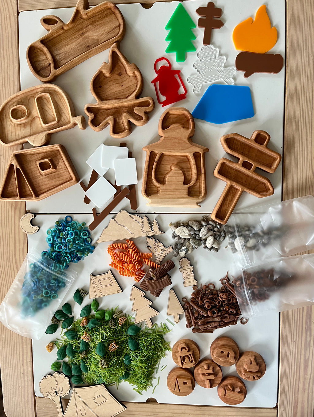 Wooden camping-themed trays with tent, camper, fire, lantern, trees, signs, play pieces, beads, and natural elements on white surface