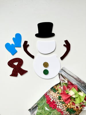 Disassembled snowman craft kit with black top hat, brown arms, blue mittens, red scarf, and glittery buttons near festive pasta in plastic bag