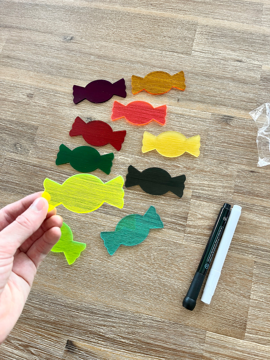 Hand holding a translucent yellow candy-shaped piece from Little Dry Erasables - Translucent Candy / Candies set, surrounded by other colorful pieces on a wooden surface with black and white dry erase markers, showcasing handcrafted homeschool education resources.