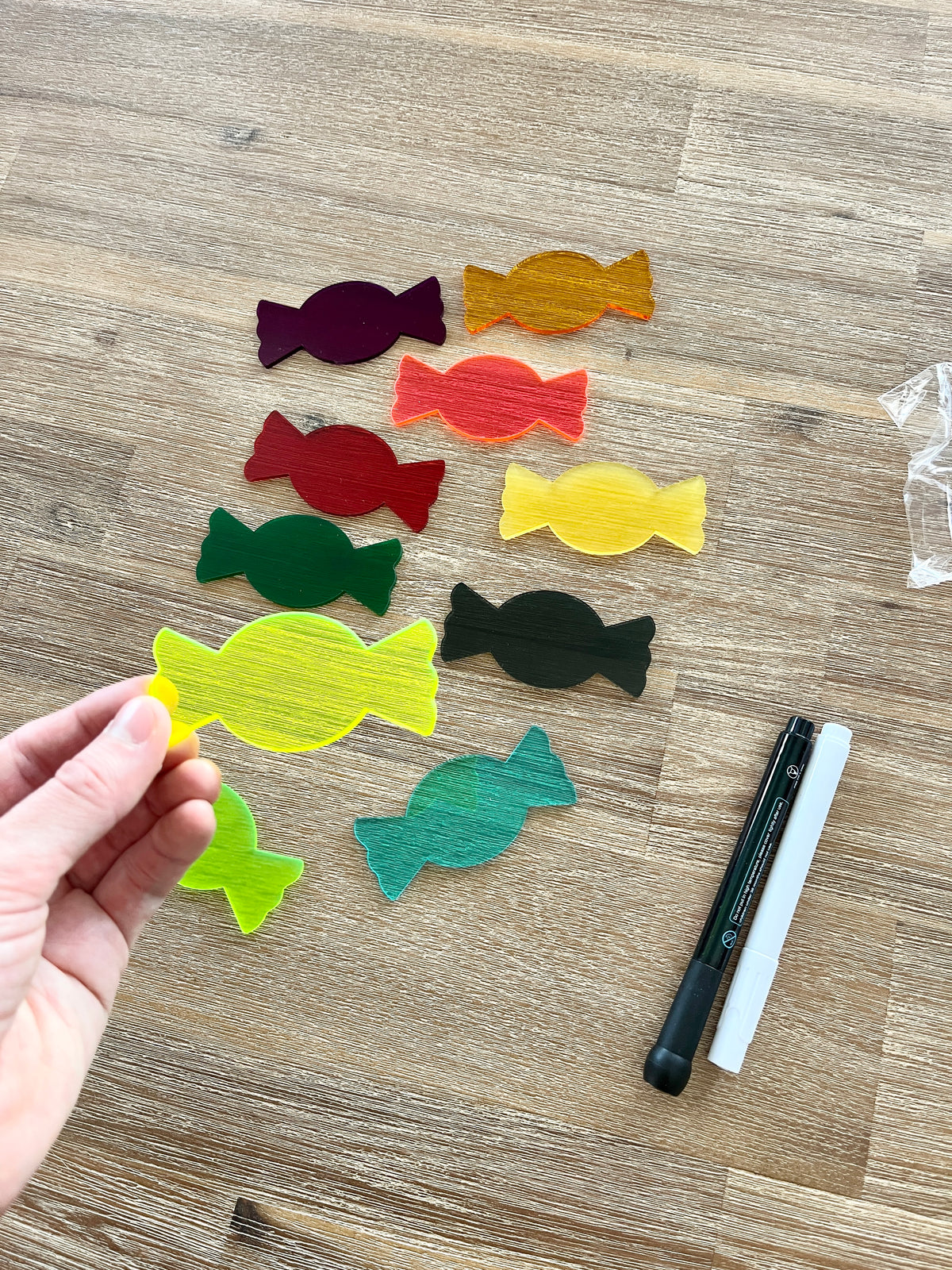Hand holding a translucent yellow candy-shaped piece from Little Dry Erasables - Translucent Candy / Candies set, surrounded by other colorful pieces on a wooden surface with black and white dry erase markers, showcasing handcrafted homeschool education resources.