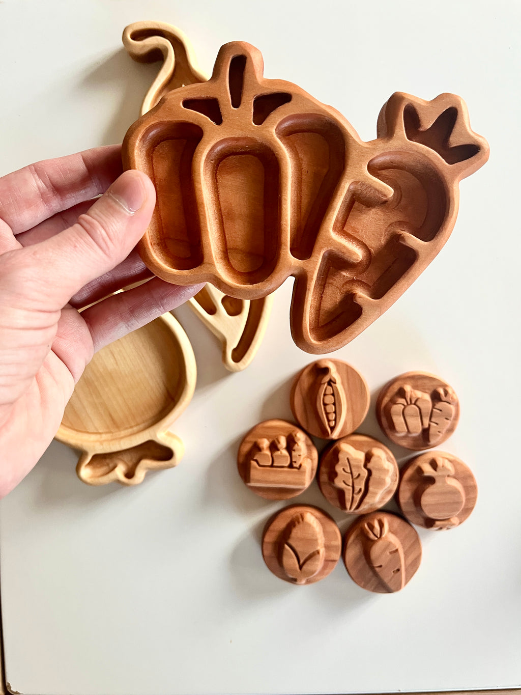 Hand holding wooden tray shaped like three carrots above round wooden stamps with vegetable carvings on white surface
