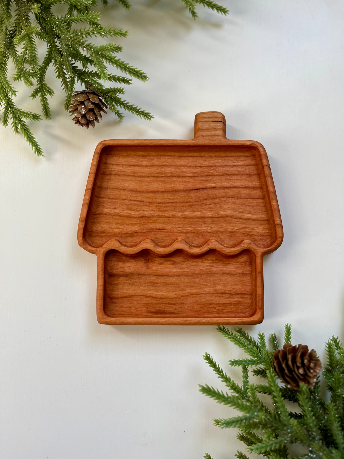 Wooden tray shaped like a house with chimney, surrounded by pine branches and pine cones on white surface