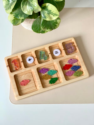 Wooden tray with colorful glittered numbers, math symbols, and assorted glittery leaf shapes on a beige surface next to a green plant
