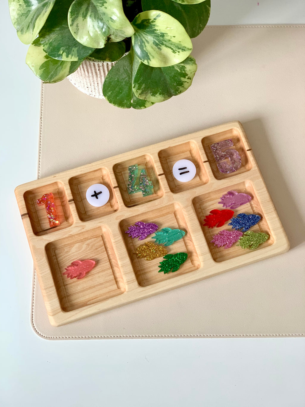 Wooden tray with colorful glittered numbers, math symbols, and assorted glittery leaf shapes on a beige surface next to a green plant