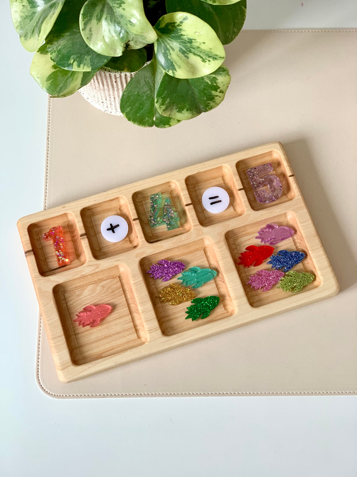 Wooden tray with colorful glittered numbers, math symbols, and assorted glittery leaf shapes on a beige surface next to a green plant