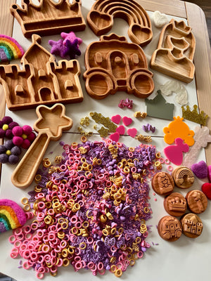 Princess-themed sensory kit with wooden trays, felt balls, colorful pasta, crowns, hearts, and rainbows on white surface