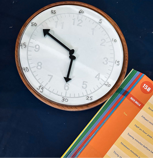 Wood-framed wall clock showing 5:50 on a dark blue surface next to colorful educational flashcards