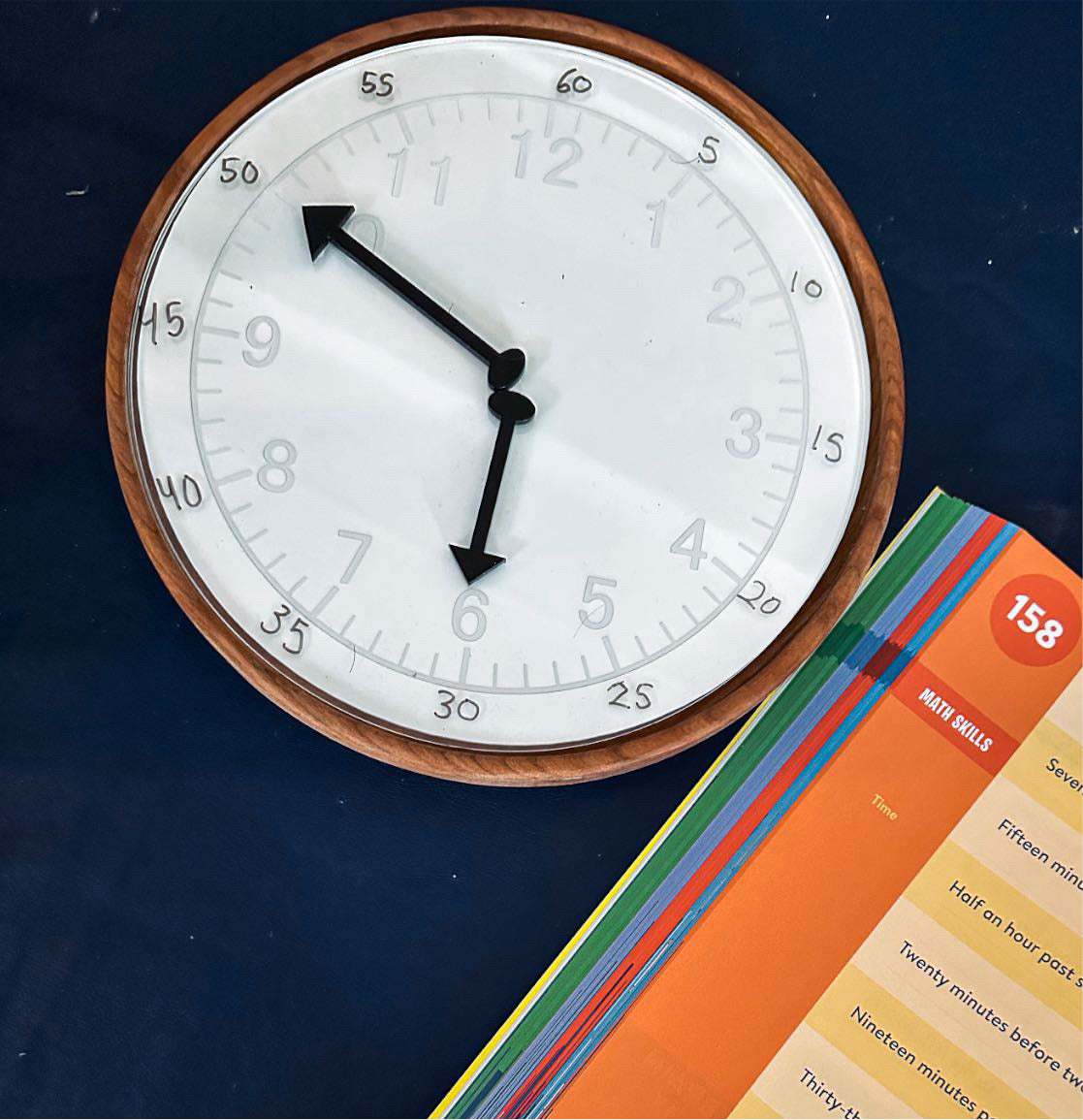 Wood-framed wall clock showing 5:50 on a dark blue surface next to colorful educational flashcards