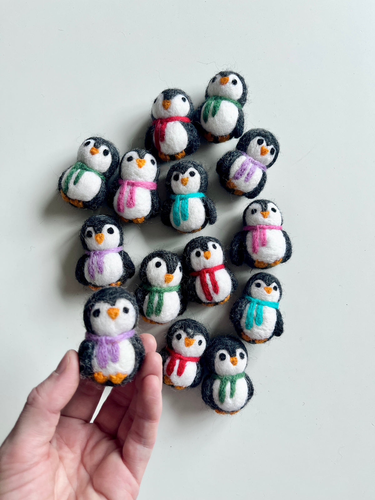 Handcrafted felt penguin figurines wearing colorful scarves arranged on white background with a hand holding one penguin