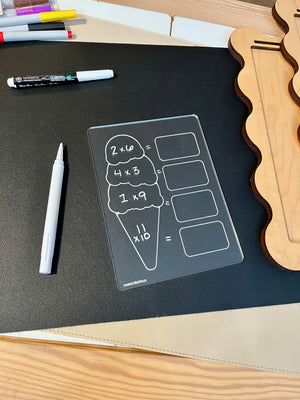 Clear acrylic math practice board shaped like ice cream with multiplication problems and white marker on black surface