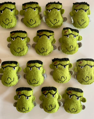 Fifteen handmade green felt Frankenstein monster face plushies with black hair and stitched scar details on beige background
