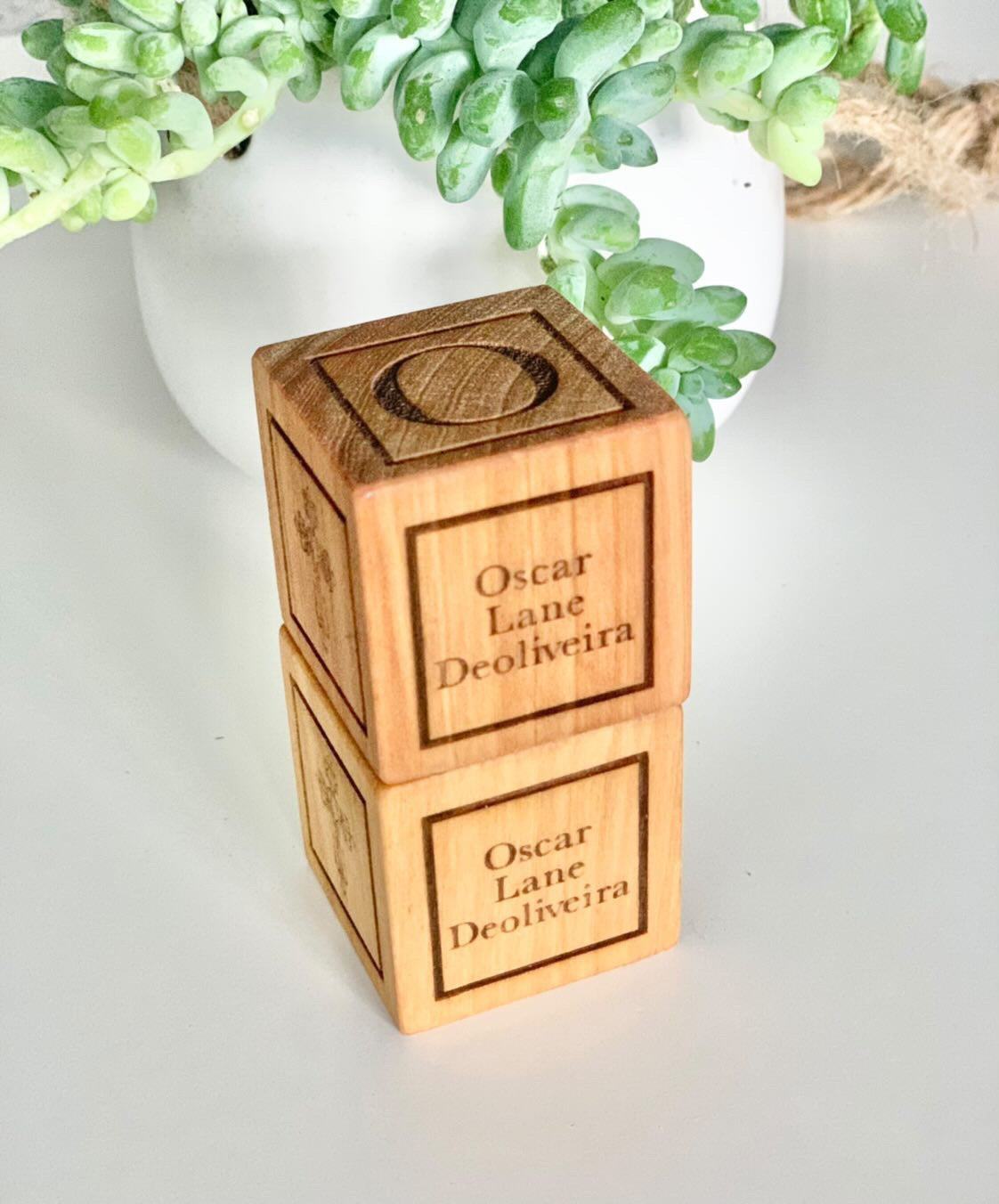 Stacked wooden baptism blocks engraved with personalized name and decorative letter on a white surface with greenery background
