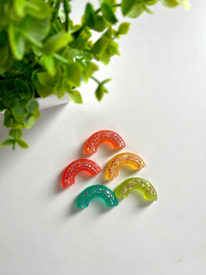 Five semi-circular resin pieces in orange, red, yellow, green, and teal with glitter on a white surface near green leaves