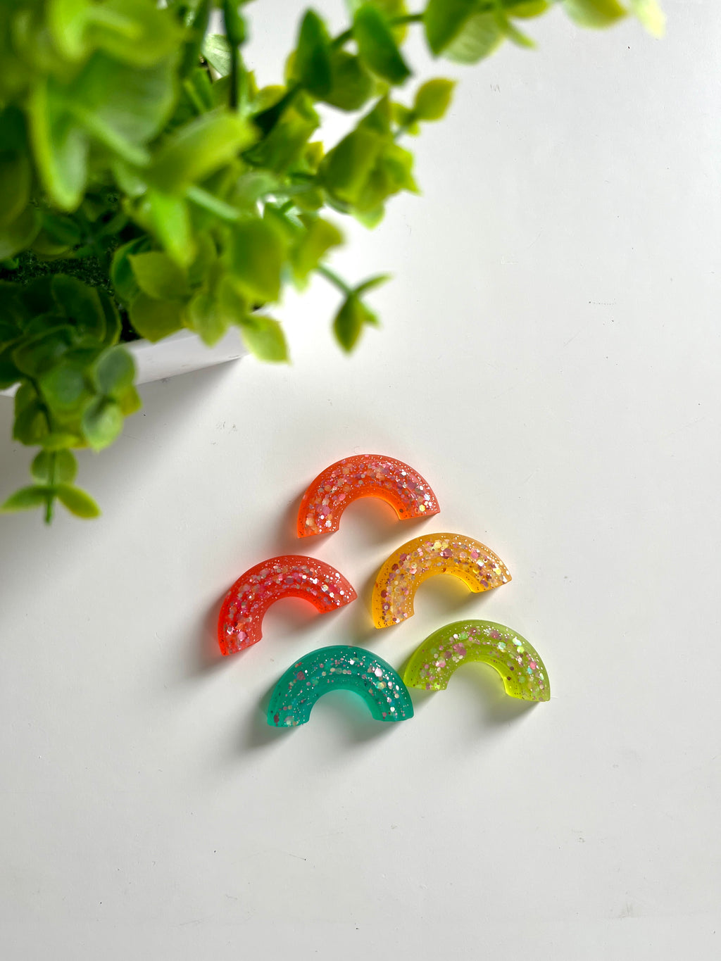 Five semi-circular resin pieces in orange, red, yellow, green, and teal with glitter on a white surface near green leaves